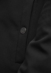 Tissu noir à texture lisse, avec un bouton métallique distinct et des coutures propres le long de la couture. Aucun motif ou accent visible.