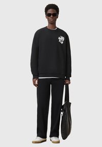Schwarzer Kapuzenpullover mit Logo auf der Brust, kombiniert mit schwarzen weit geschnittenen Hosen. Das Model hält eine schwarze Ledertasche, trägt schwarze Sonnenbrillen und weiße Sneaker.