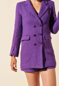Blazer texturé violet avec une coupe ajustée, boutons double-breasted et short assorti. Les détails notables incluent des poches à soufflet et des revers angulaires.