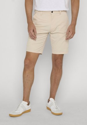 Man draagt beige knielange shorts en witte sneakers, staand tegen een effen lichtgrijze achtergrond.