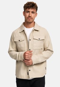 Beige Jacke aus weichem Stoff, mit durchgehender Knopfleiste, zwei Brusttaschen, langen Ärmeln und schwarzen Knopfakzenten.