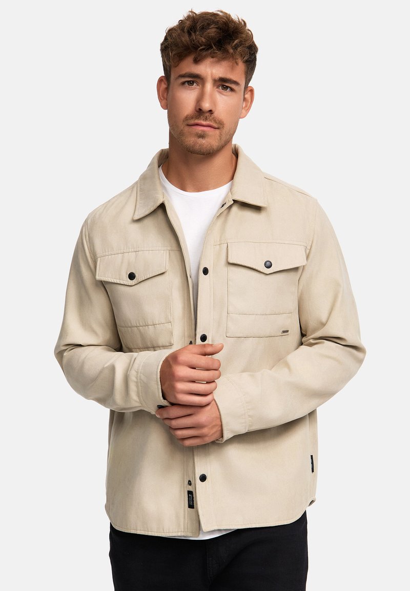 Beige Jacke aus weichem Stoff, mit durchgehender Knopfleiste, zwei Brusttaschen, langen Ärmeln und schwarzen Knopfakzenten.