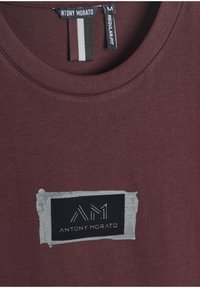 Camiseta de algodón burdeos con cuello redondo, que cuenta con un parche rectangular gris con el logotipo "ANTONY MORATO" en letras negras y blancas.