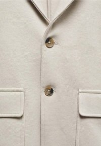 Beige texturerad jacka med två framfickor och bruna knappar. Tyget har ett subtilt vävmönster och en notch lapel-design.