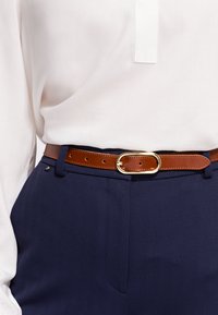 Valkoinen paita, joka on puettu sinisiin haarukkatakkihousuihin ruskealla nahkavyöllä, jossa on kultainen ovaali solki.