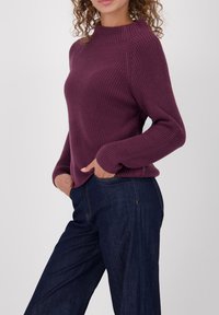 Gebreide trui in diep paars met een geribbelde textuur, voorzien van een hoge halslijn en lange mouwen. Gecombineerd met donkerblauwe denim broek.