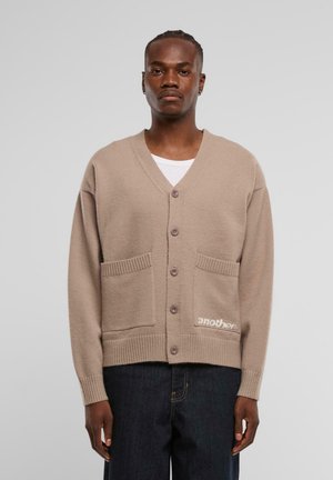 Beige gestrickte Strickjacke mit V-Ausschnitt, Knöpfen, zwei vorderen Taschen und gerippten Bündchen; hat "another" auf der unteren rechten Seite gestickt.