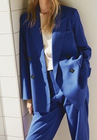 Blå dubbelknäppt kavaj med stora knappar och lösa ärmar, i kombination med matchande byxor och en vit skjorta under. Slätt tyg.