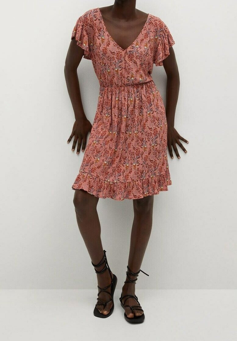 Femme en robe fleurie rose mi-longue avec manches volantées et sandales plates noires à lacets, debout devant un fond blanc uni.
