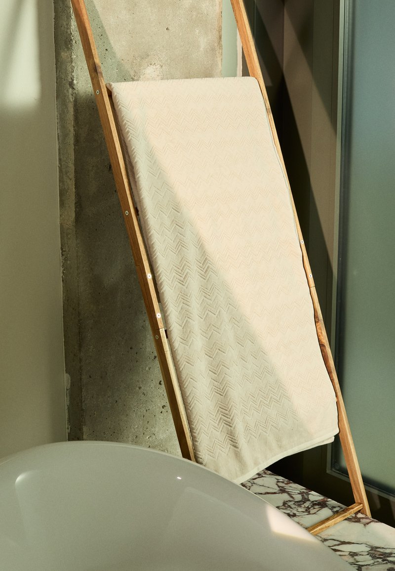 Beige színű törölköző, halszálka mintával, egy fából készült keretre terítve, betonalapú fal és márvány felület előtt.