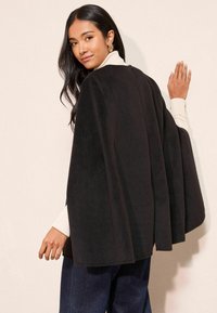 Černý fleecový plášť s volným designem, s vysokým límcem a dlouhými rukávy, nošený přes světle barevný rolák a džíny.