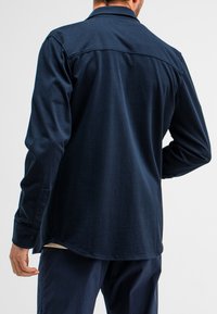 Dunkelblaues Langarmshirt mit klassischem Kragen, geknöpften Manschetten, abgerundetem Saum und glatter Stoffoberfläche. Details sind am Rücken sichtbar.