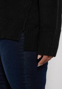 Czarny sweter z dzianiny z prążkowanymi mankietami i dołem, z bocznymi rozcięciami dla swobody ruchów, noszony na ciemnych dżinsach.