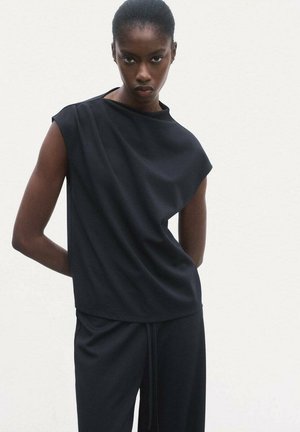 Black short-sleeve top with asymmetrical design, soft fabric, relaxed fit, and slightly draped back. Paired with black pants, minimal style.