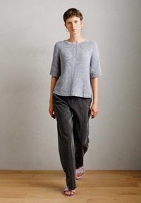 Donna con i capelli corti che cammina all'interno, indossando un maglione grigio chiaro a maniche corte, pantaloni scuri, orecchini a cerchio argentati e sandali viola.