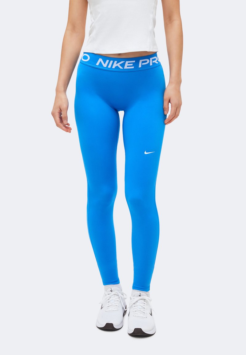Άτομο που φοράει φωτεινά μπλε κολάν Nike Pro με λευκό λογότυπο Nike, λευκό αθλητικό τοπ και λευκά αθλητικά παπούτσια στέκεται μπροστά σε απλό φόντο.
