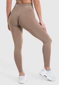 Bruine hooggewaisted leggings van stretchy stof, met platte naden en een logo op de tailleband, gecombineerd met witte sportschoenen.