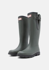 Botas de chuva de borracha verdes com bico arredondado, superfície texturizada e sola cinza. Apresenta uma fivela e um rótulo de marca perto da abertura.