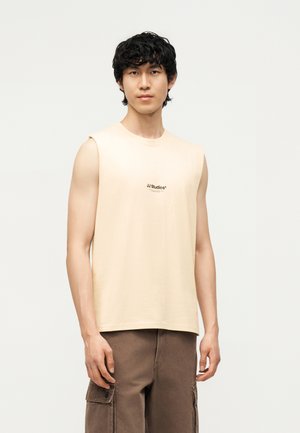 Ung mann med krøllete svart hår, iført en beige ermeløs skjorte og brune cargobukser, stående mot en ensfarget lys bakgrunn.