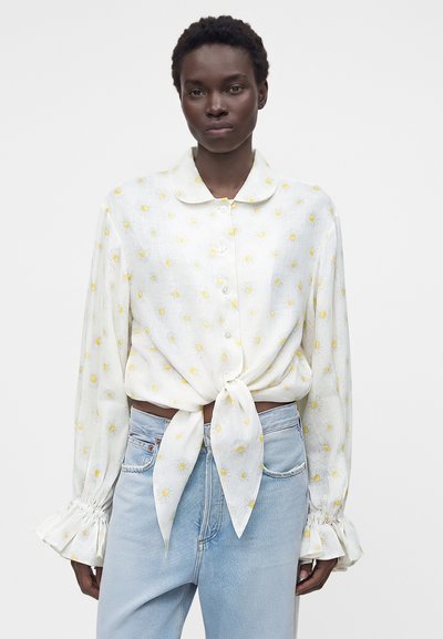 Γυναίκα με λευκό πουκάμισο με κίτρινα σχέδια με μαργαρίτες, δεμένο στη μέση, συνδυασμένο με ανοιχτού μπλε τζιν σε απλό φόντο.