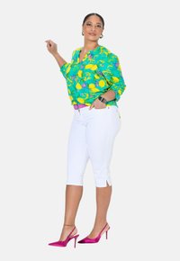 Blusa verde brillante con motivo di limoni gialli e fiori viola, pantaloni bianchi fino al ginocchio e tacchi a spillo rosa. Orecchini a cerchio d'argento e bracciale.