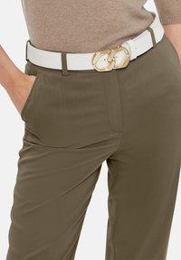 Olijfgroene broek met een gladde textuur, voorzien van zijzakken. Gecombineerd met een witte leren riem, versierd met een gouden kettinggesp.