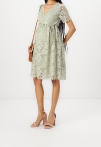 Valkoinen pitsimekko pehmeässä vihreässä, jossa on V-pääntie, lyhyet hihat, vyötärönauha ja aaltoileva helma. Yhdistetty beigen väristen paksupohjaisten sandaalien kanssa.