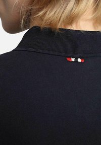 Camisa polo negra con cuello acanalado, que presenta un pequeño detalle bordado en tri-color en rojo, blanco y azul en la parte posterior del cuello.