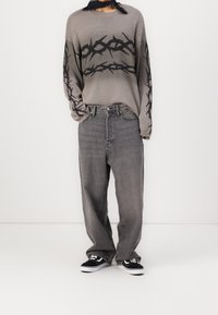 Šedý pletený svetr s černým grafickým motivem trnu, zkombinovaný s volnými šedými džínami a černo-bílými teniskami. Celkově ležérní outfit.