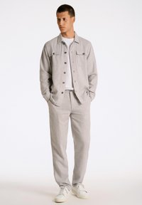 Ljust grå skjorta med knappar och två framfickor, parat med ljusgrå drivrembyxor, bärs med vita sneakrar. Slät textur, avslappnad design.