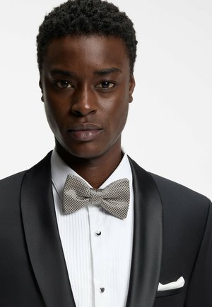 Smoking nero con rever in raso, camicia bianca plissettata e papillon a quadri grigio. Dettagli notevoli includono un fazzoletto da taschino e gemelli.