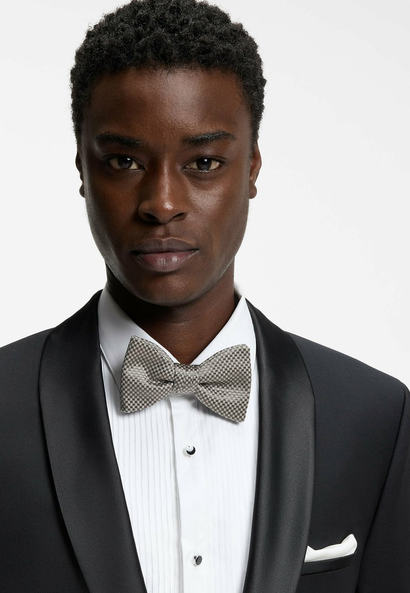 Smoking nero con rever in raso, camicia bianca plissettata e papillon a quadri grigio. Dettagli notevoli includono un fazzoletto da taschino e gemelli.