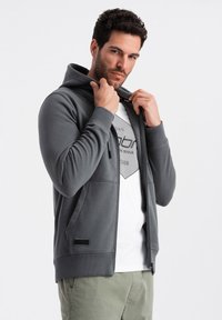 Grau zip-up Hoodie aus weichem Stoff mit Kapuze, vorderen Taschen, gerippten Bündchen und einem schwarzen Akzentlabel am Saum. Getragen über einem weißen T-Shirt mit Grafik.
