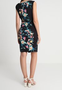 Schwarzes Blumenkleid mit kurzen Ärmeln, das ein buntes Blumenmuster aufweist. Das Model trägt weiße, schnürbare High-Heels.