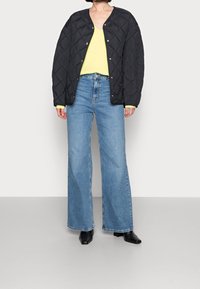 Svart vadderad jacka med knappstängning, gul långärmad skjorta, vida blå denimjeans och svarta spetsiga ankelstövlar.
