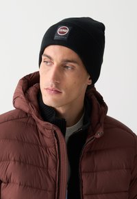Svart stickad mössa med en fyrkantig logotagg; bärs med en bordeauxfärgad puffjacka med dragkedja framtill och texturerad krage under.