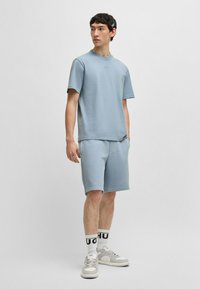 T-shirt en coton bleu et shorts assortis, coupe décontractée, associés à des baskets blanches et des chaussettes noires et blanches contrastantes, se tenant devant un fond uni.