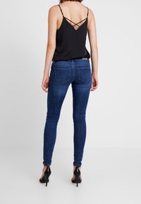 Dunkelblaue Skinny-Jeans mit fünf Taschen, leichtem Verblassen und genähten Details. Kombiniert mit einem schwarzen Camisole mit überkreuzten Trägern.