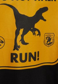 Černé a žluté grafické tričko s potiskem siluety dinosaura, odvážným textem "NECHOĎTE! BĚŽTE!" a detailem odznaku služby pro ochranu přírody.