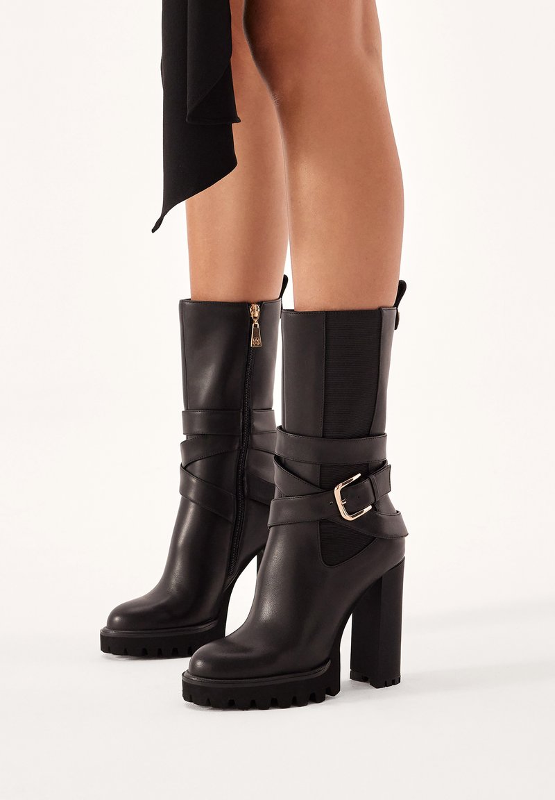 Botas de tobillo de cuero negro con un tacón grueso, cremallera lateral, paneles elásticos y una correa decorativa con hebilla. Suela texturizada para mayor agarre.