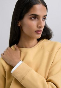 Żółta bluza z ribbed cuffs oraz białą podszewką, noszona przez osobę z długimi ciemnymi włosami, częściowo widoczna ręka chwytająca materiał.