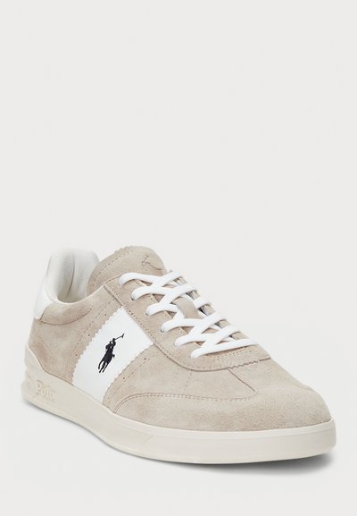 Beige suede sneaker with white accents and laces. Features a logo on the side and a rubber sole with textured detailing.