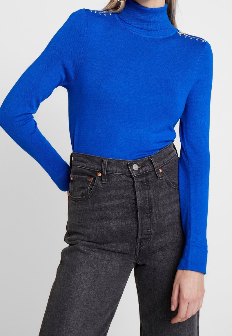 Blauer Rollkragenpullover mit langen Ärmeln, verziert mit metallischen Nieten an den Schultern, kombiniert mit hochgeschnallten schwarzen Jeans.