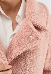 Roze gestructureerde jas van pluizig materiaal, met een notch-kraag en een gouden accentknop. Een knoopshirt is zichtbaar eronder.