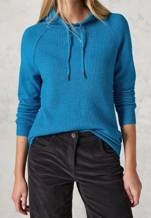 Femme portant un sweat à capuche en tricot bleu texturé avec cordons et un pantalon en velours côtelé noir taille haute, debout devant un fond clair et neutre.