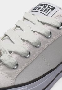 Λευκό πάνινο παπούτσι Converse All Star με λευκά κορδόνια που περνούν μέσα από μεταλλικά παπούτσια, παρουσιάζοντας την ετικέτα με το λογότυπο στη γλώσσα του παπουτσιού.