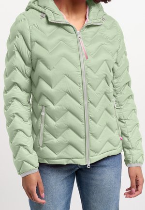 Personne portant une veste matelassée à capuche vert clair avec fermeture éclair avant et poches zippées, associée à un jean bleu.