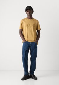 Yellow cotton t-shirt with "BOSS" printed in a darker shade, paired with blue denim jeans and black lace-up sneakers.