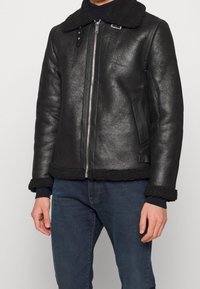 Giacca in shearling nera con zip frontale, ampio colletto e finitura in pelle effetto strutturato. Presenta un orlo e polsini a costine, con tasche laterali.