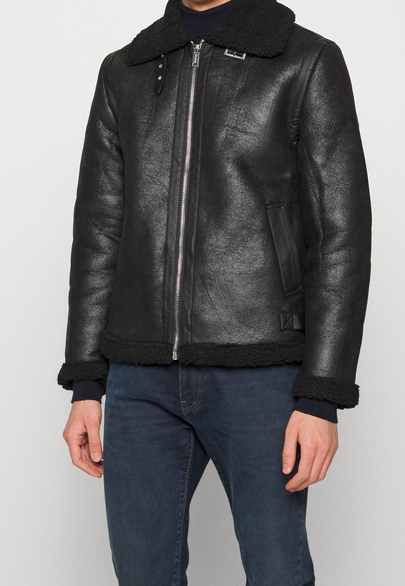 Giacca in shearling nera con zip frontale, ampio colletto e finitura in pelle effetto strutturato. Presenta un orlo e polsini a costine, con tasche laterali.